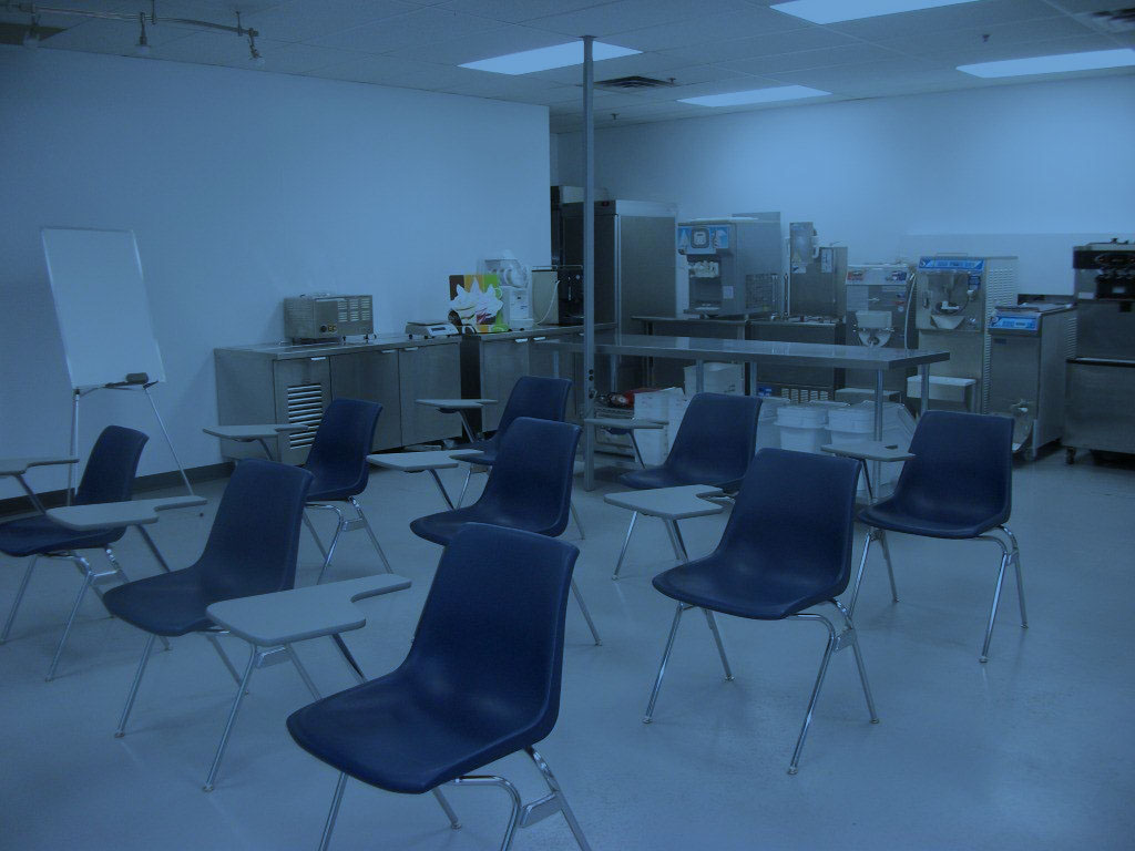 Empty classroom-style kitchen with rows of blue chairs with desks, stainless steel counters and large professional machines in the background, desaturated monochrome film photograph with visible grain