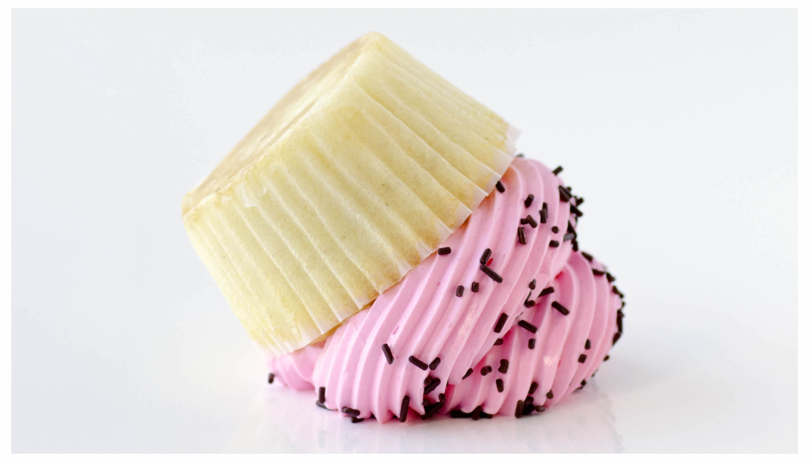 A small elegant pastry with pink frosting and chocolate sprinkles on a plain white background, bright diffused daylight, grainy film texture.